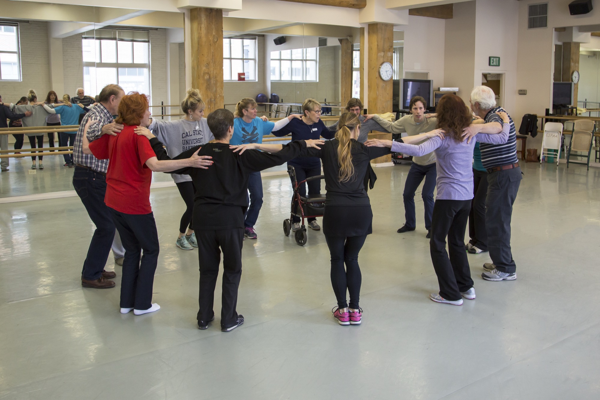 A circle of dancers, hands on each other's shoulders, in a mirrored studio